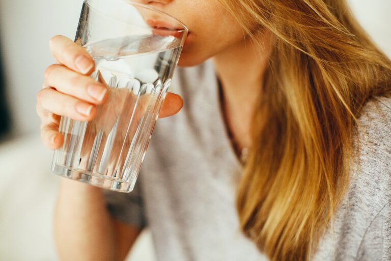 Eine Frau mit blonden Haaren und einem grauen T-Shirt trinkt ein großes Glas mit Wasser