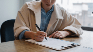 Eine Frau sitzt an einem Tisch und schreibt auf ein Blatt auf einem Klemmbrett