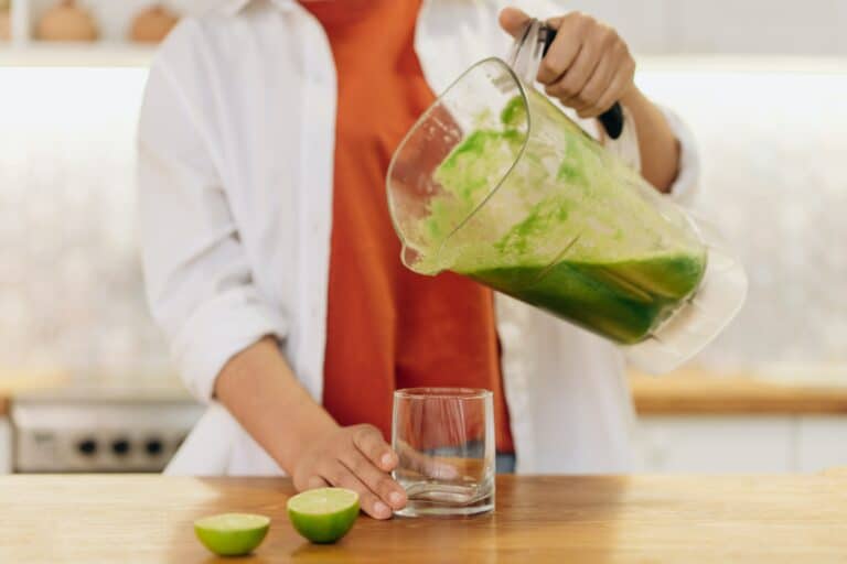 Eine Person mit einem orangefarbenen T-Shirt und einem weißen Hemd steht in einer Küche und schenkt sich einen grünen Smoothie in ein Glas.