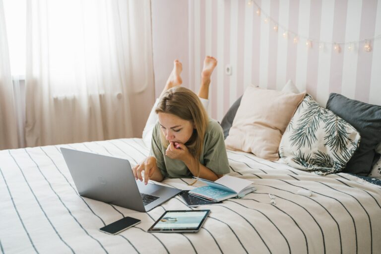 Eine blonde Frau mit einem grünen T-Shirt und einer weißen Hose liegt auf einem Bett und arbeitet am Laptop. Neben ihr liegen ein Notizbuch, ein Smartphone und ein Tablet.