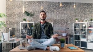 Mann sitzt in einer Yogaposition auf dem Schreibtisch.